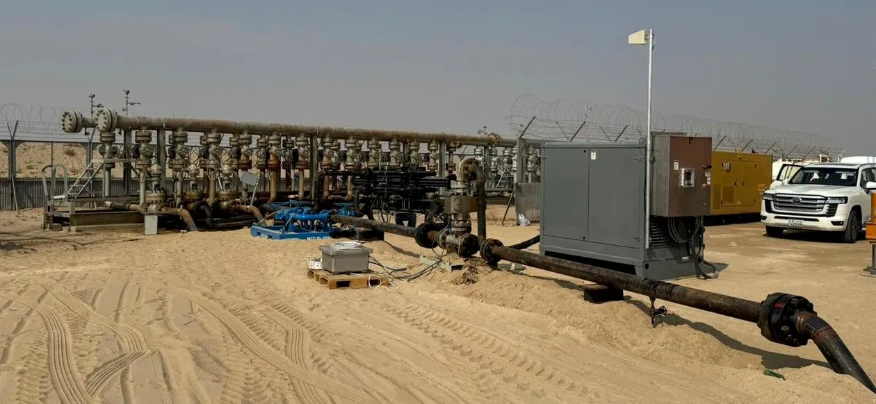 Wellhead manifold with a row of valves on a sandy site, a grey pump enclosure and a flowline in the foreground