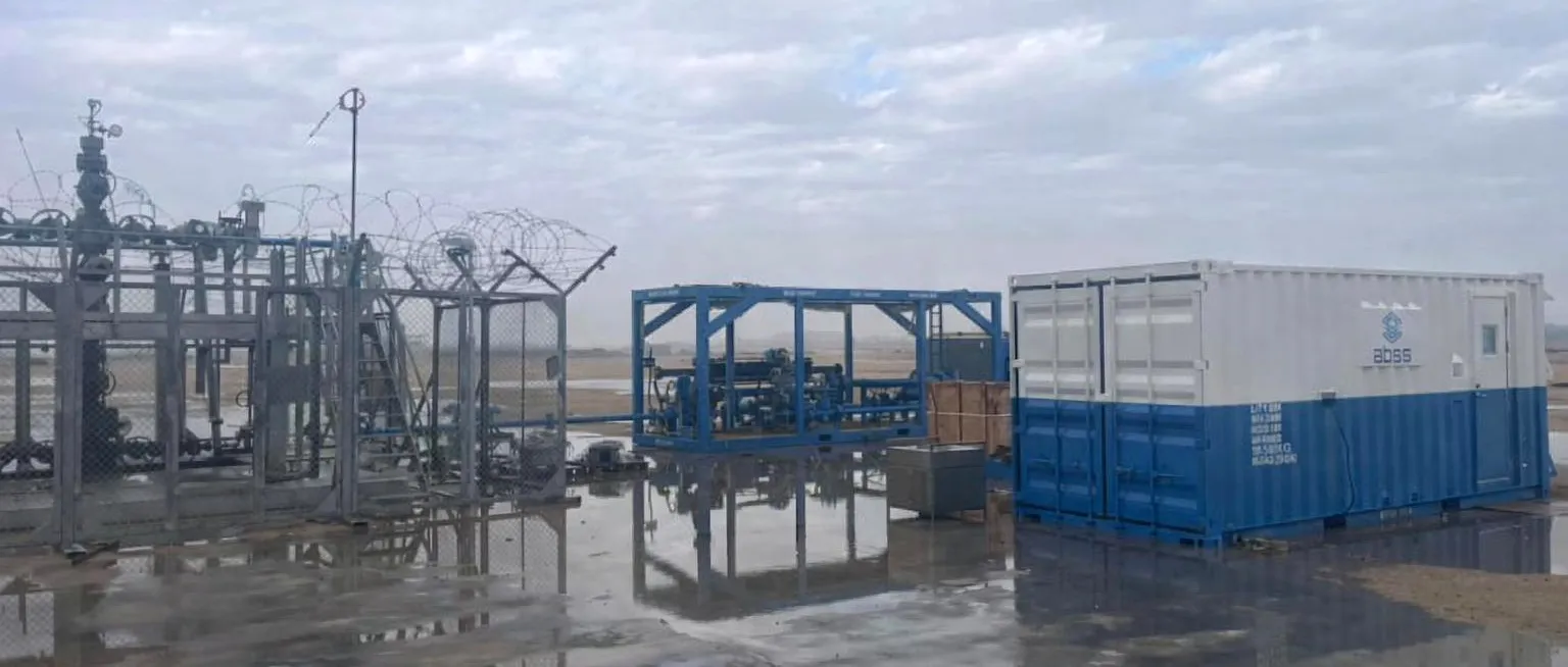 BoostFlow skid and a blue-and-white ABSS equipment container beside a fenced wellhead on a wet, overcast wellsite